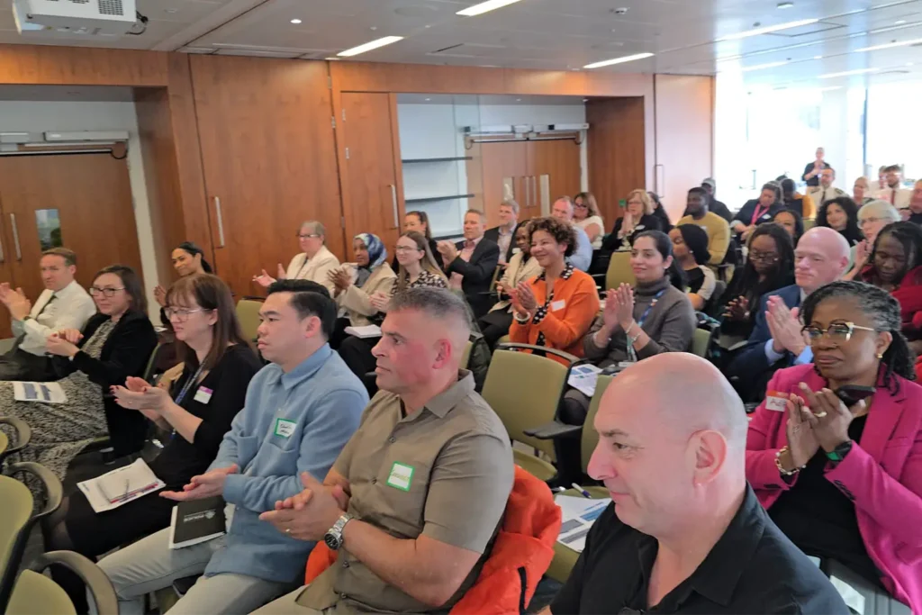 Audience of seated people applauding at an indoor event, with a large group of attendees in rows behind them.