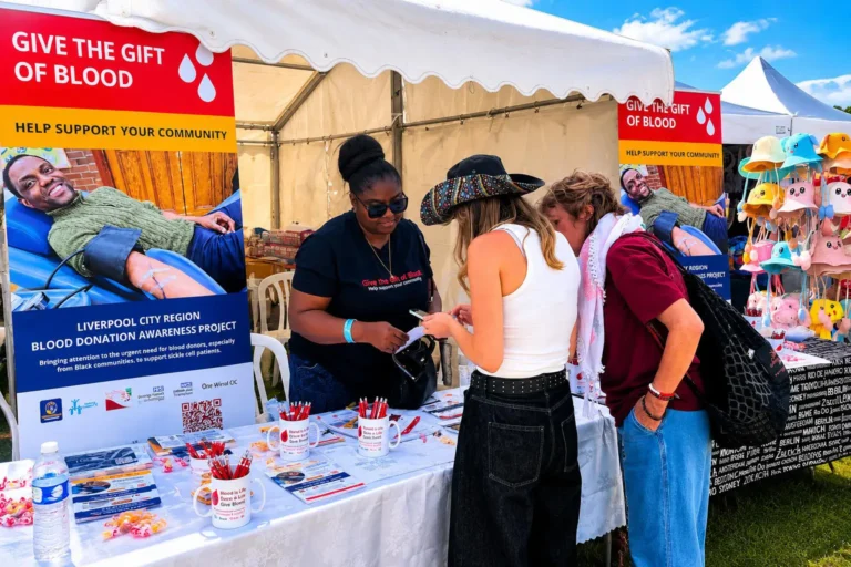 Outreach stall at an outdoor event: a team member and two visitors talk at a table of leaflets, with 'Give the gift of blood' banners behind