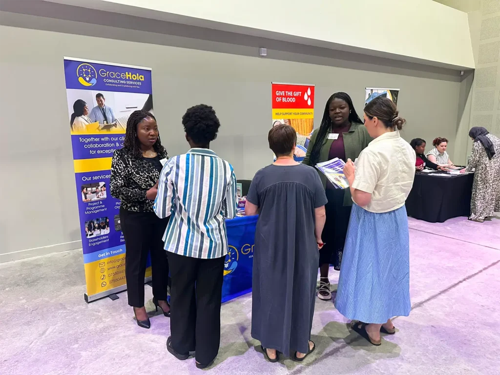 Four people in conversation at an indoor community event, beside a GraceHola banner and a 'Give the gift of blood' banner