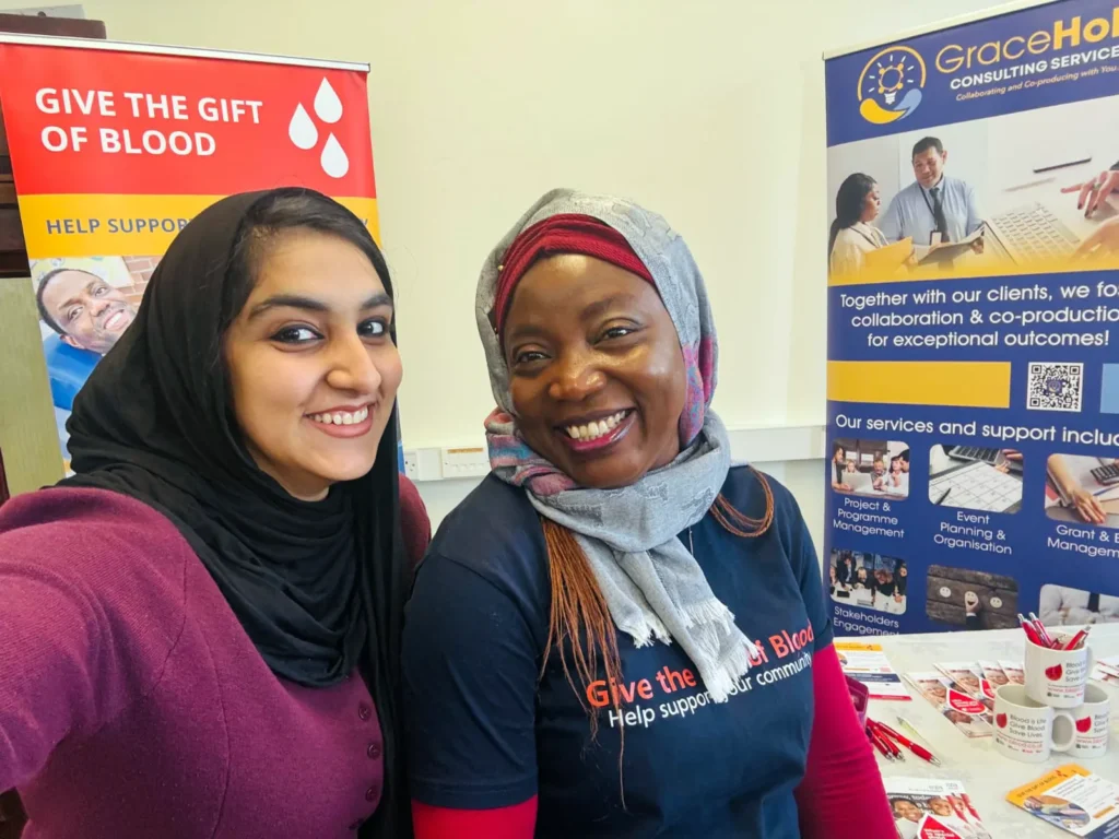 Two smiling women taking a close-up photo together in front of a 'Give the gift of blood' banner and a GraceHola banner