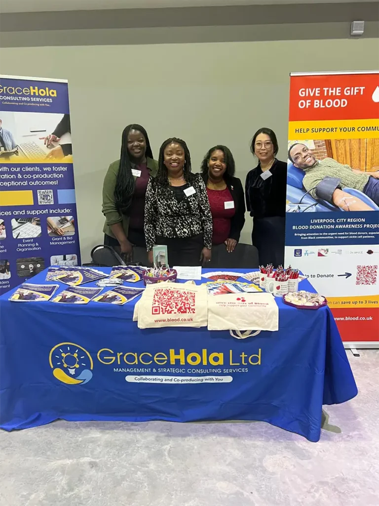 Four women standing behind a blue GraceHola table at an indoor event, with banners on either side and leaflets on the table