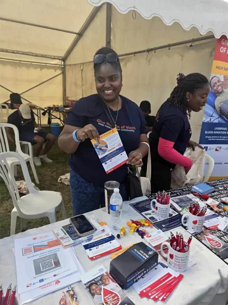 A smiling volunteer holds up a campaign leaflet at a stall table covered with leaflets, pens and other materials